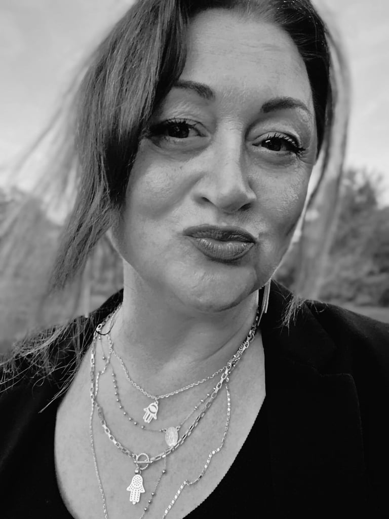 Black and white portrait of a woman wearing layered silver necklaces with charm pendants, looking directly at camera with a serious expression.