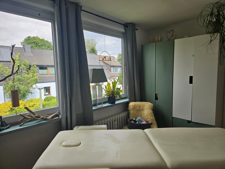 Bright bedroom with white massage table, green wardrobe, large windows overlooking residential neighborhood, and potted plants