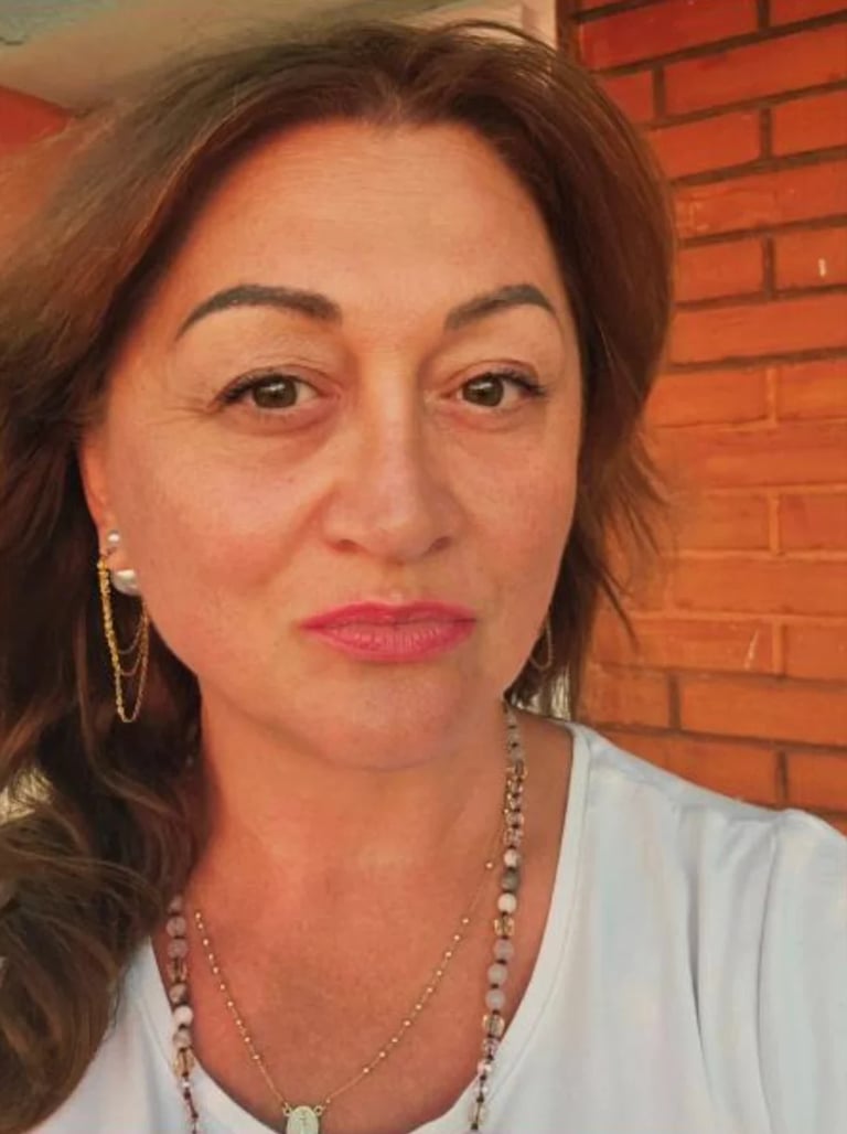 Woman with brown wavy hair, pink lipstick, and pearl necklace against a red brick background