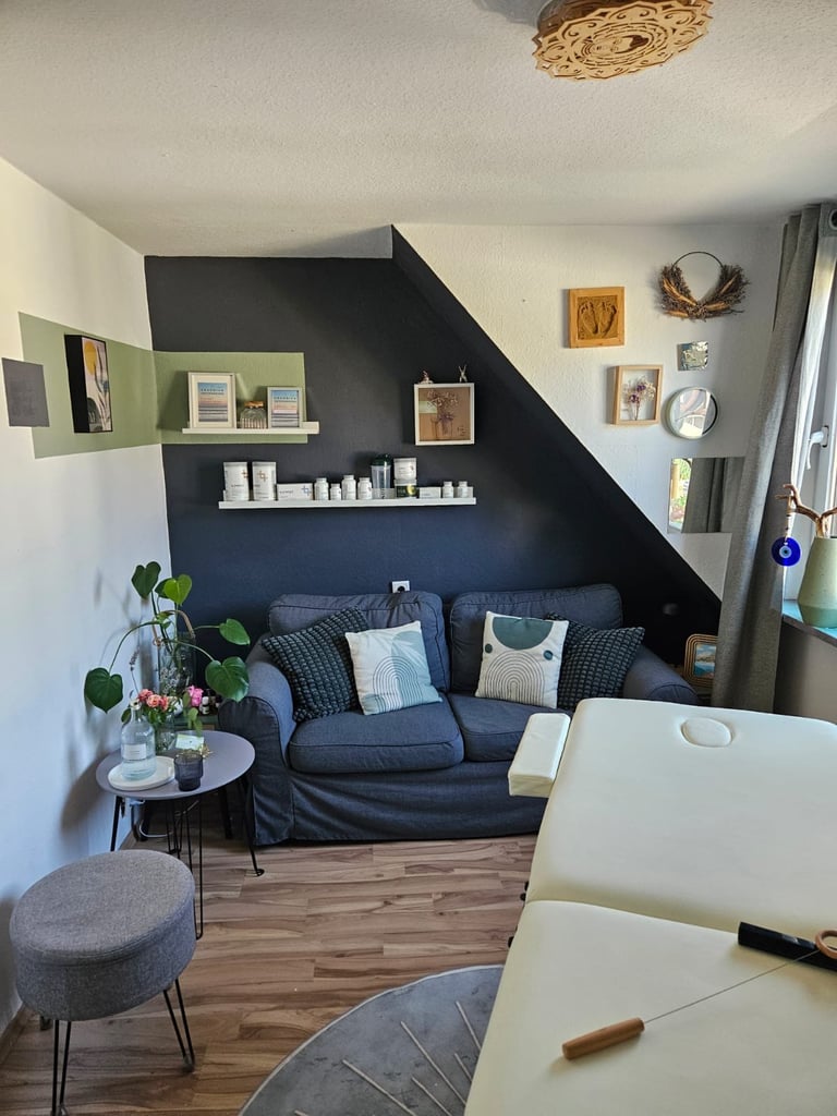 Cozy home office nook with dark blue sloped wall, navy sofa, floating shelves, small side table with flowers, and wooden desk in foreground