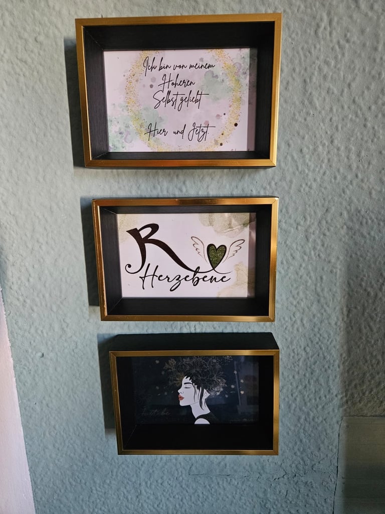 Three framed artworks hanging vertically on a light blue wall with gold-trimmed wooden frames. Top frame shows handwritten text with watercolor design, middle frame displays Herzscheine text with a heart design, and bottom frame shows a black and white portrait image.